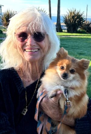 Smiling woman with long white hair and sunglasses holds a small Pomeranian dog, set against a sunny park backdrop with palm trees.